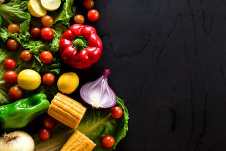 Vegetables arranged on black table with empty space on right. Black surface with green pepper, corn, cherry tomatoes, lemons, onions and lettuce.の写真素材