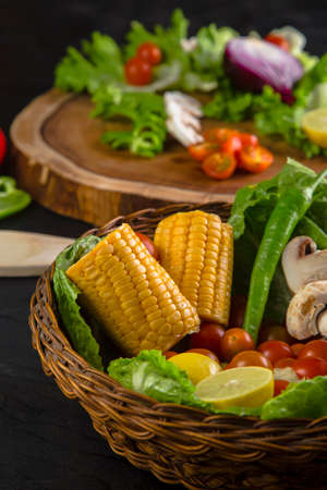 Cropped rattan bowl with vegetables on black table. Wicker basket with corn and greens in front of wooden slice with mushrooms, lettuce and tomatoes.の写真素材