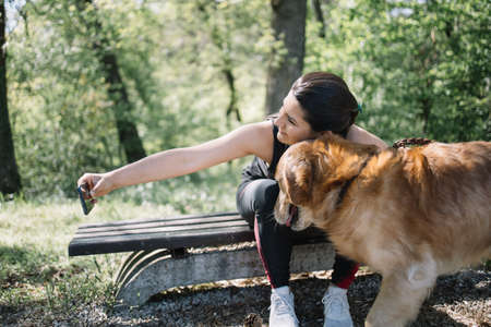 Smiling girl taking selfie with her dog. Side view of girl sitting on bench and taking selfie while hugging dog.の写真素材