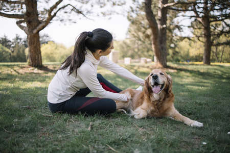 Lady and dog enjoying sunny weather in park. Woman in sport outfit petting her retriever dog while sitting on meadow in park.の写真素材