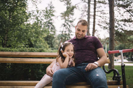 Father and daughter sitting on bench in city park. Father hugging his daughter while sitting on bench in park.の写真素材