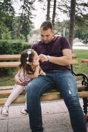 Man pinching little girls cheek while resting on bench. Father and his little daughter sitting on wooden bench in city park and playing.の写真素材