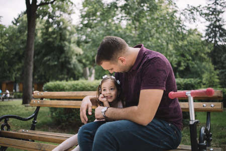 Little girl cuddling with her father in city garden. Man hugging little girl while resting on bench in park with trees in background.の写真素材