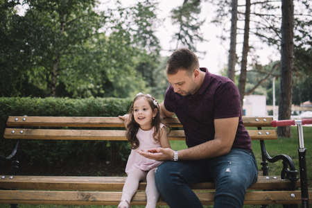 Girl with flower crown sitting on bench next to her father. Young father holding hand of his daughter while sitting on bench.の写真素材