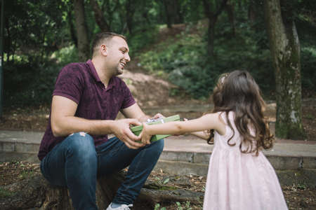 Dad and small girl holding present in park. Father sitting on stump in park giving present to his...