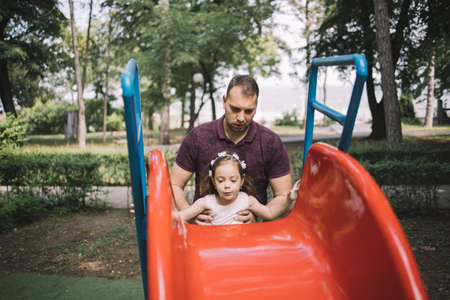 Young father holding her daughter behind slide. Man holding little girl to climb slide in playground.の写真素材