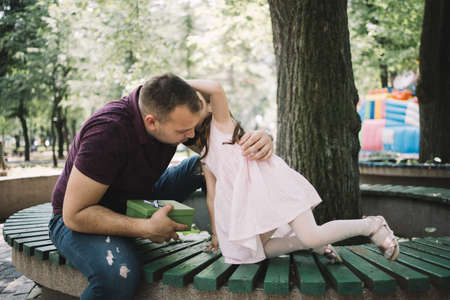 Daughter whispering to her daddy who is holding gift. Young father holding present while sitting on bench with his child.の写真素材