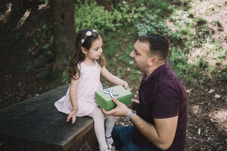 Father receiving gift box from her daughter outdoor. Daughter giving present to her father who is sitting on bench in park.の写真素材