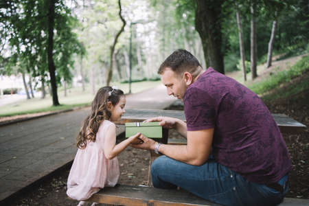 Father and little girl exchanging presents in nature. Side view of daughter and father sitting on bench while holding green gift box.の写真素材