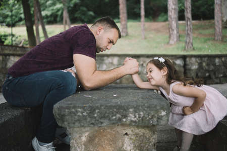 Young man and little child arm wrestling. Father and his little daughter wrestling with arms over stone table in park.の写真素材