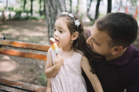 Daughter eating ice cream while sitting on bench with her father. Cute little girl with headband eating ice cream while her dad is looking at her.の写真素材