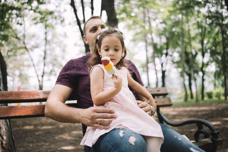 Cute little girl with headband eating ice cream in nature. Daughter looking at her ice cream while sitting in her fathers lap in forest.の写真素材
