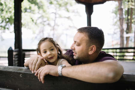 Portrait of dad hugging his daughter in wooden gazebo. Man cuddling little girl while leaning on wooden fence in nature.の写真素材