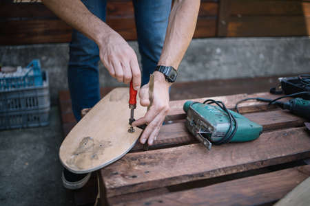 Mans hands making skateboard and using screwdriver. Cropped man assembling skateboard on table with...