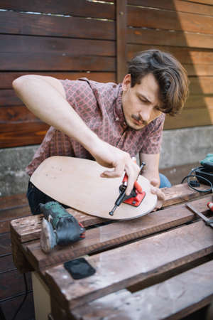 Portrait of man mounting skateboard trucks on wooden deck. Focused man putting together skateboard...