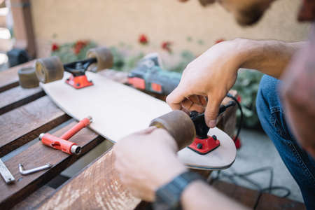 Cropped man screwing skateboard wheel to board in backyard. Close-up view of male hands fitting...