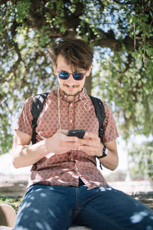 Portrait of smiling man using phone under tree. Close-up view of guy with sunglasses using his...