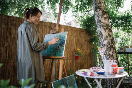 Young beautiful woman painting a picture on canvas. Happy female artist painting landscape while...