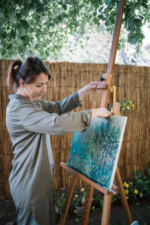 Girl getting canvas from tripod in garden. Womans hand holding painting and canvas stand while standing in backyard with flowers and plants.の写真素材