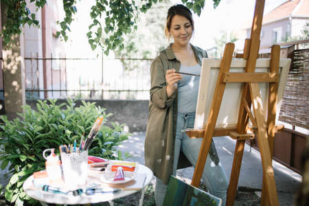 Female artist using paint brush while painting in backyard. Beautiful woman painting while standing in garden next to table with brushes and paint tubes.の写真素材
