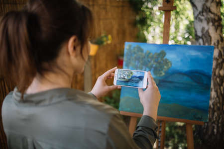 Woman taking photo of painting on canvas stand in garden. Side view of woman artist taking photo of painted canvas on tripod placed in yard.の写真素材