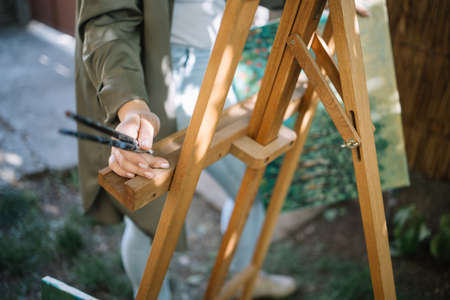 Close-up view of female hand holding paint brushes over tripod. Cropped woman holding paint brushes while standing in garden with tripod.の写真素材