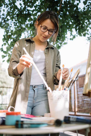Young woman taking paint brush from pot with brushes. Artist choosing paint brush and preparing for...