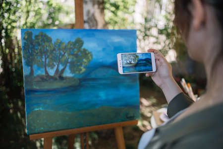 Backview of woman taking photo of painted canvas. Cropped girl holding phone and taking photo of painting on tripod in nature.の写真素材