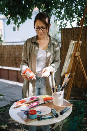 Womens artist preparing for painting in garden. Pretty woman wearing glasses squeezing paint tube in outdoor workshop.の写真素材
