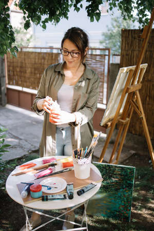 Beautiful female artist standing in outdoor studio. Pretty woman wearing glasses holding paint tube over table with paint brushes and paint tubes.の写真素材