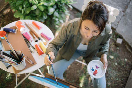 High angle view of female painter painting picture on canvas. Portrait of lady painter with paintbrush sitting in yard and painting.の写真素材