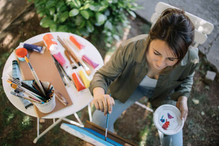 High angle view of female painter painting picture on canvas. Portrait of lady painter with paintbrush sitting in yard and painting.の写真素材