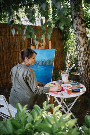 Female artist drawing picture in yard with tree and plants. Back view of woman painter painting landscape picture in outdoor garden studio.の写真素材
