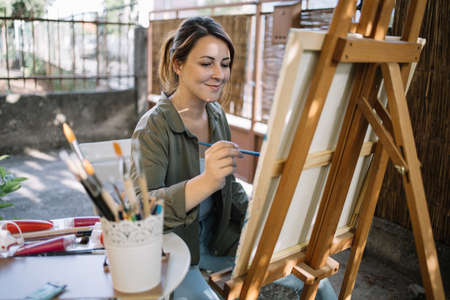 Smiling painter drawing picture using aquarelle paint and paintbrush. Calm female artist painting while sitting in backyard art studio.の写真素材