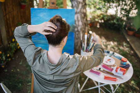 Back of woman making hairstyle while sitting in garden art studio. Back view of woman making hair bun using paintbrush in a backyard with painting.の写真素材