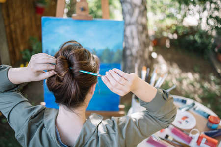 Woman making hair bun using paintbrush in yard. Close-up view of female artist making hair bun with paintbrush in front of tripod with painted canvas.の写真素材