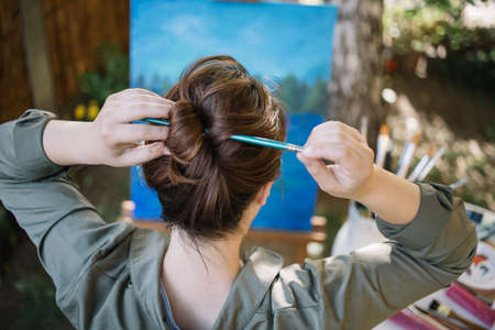 Close-up view of hair bun made with paintbrush. Woman using paintbrush to make hair bun while sitting in garden in front of painted canvas.の写真素材