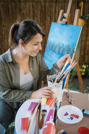 High angle portrait of woman taking paintbrush in outdoor studio. Female painter holding pot with paint brushes while sitting next to painted canvas on stand.の写真素材