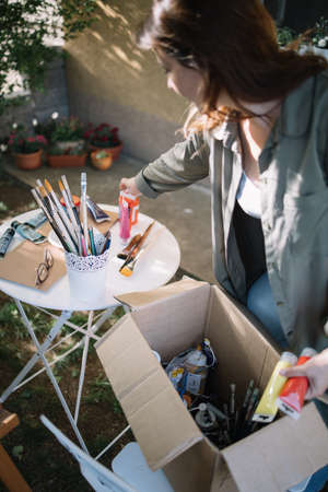 Brunette woman taking paint tubes from paper box. Woman painter picking colorful paint tubes from...