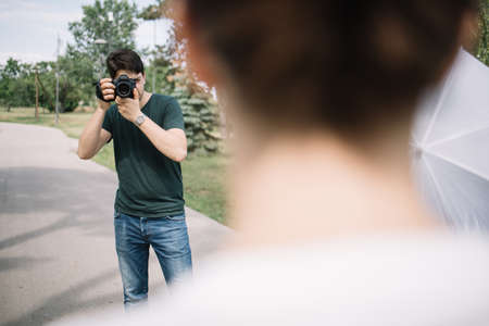 Photographer looking through camera viewfinder behind blurred woman. Photographer taking photos of girl posing on street in park.の写真素材
