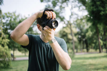 Front view of man taking photos in nature. Portrait of photographer looking through camera viewfinder and taking pictures while standing in park.の写真素材