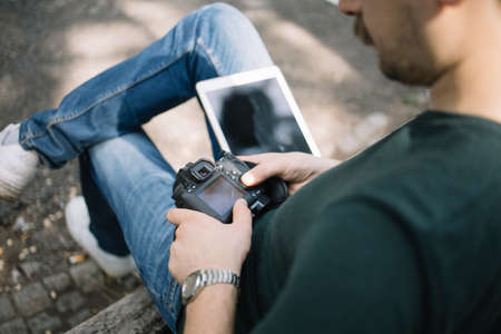 Man sitting on bench with tablet and professional camera. High angle view of photographer holding camera and having tablet in his lap while sitting on seat outdoor.の写真素材