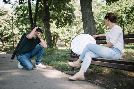 Squatting man with professional camera photo shooting female model. Side view of girl sitting on bench with reflector while photographer is taking photos of her.の写真素材