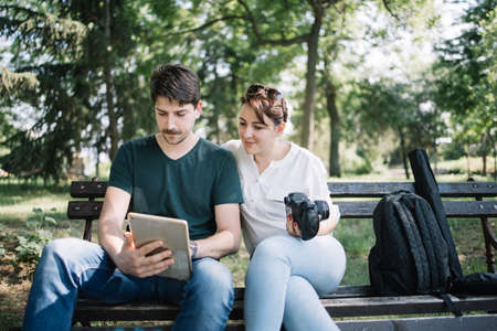 Happy couple sitting on bench in park and looking at tablet. Female model and male photographer...