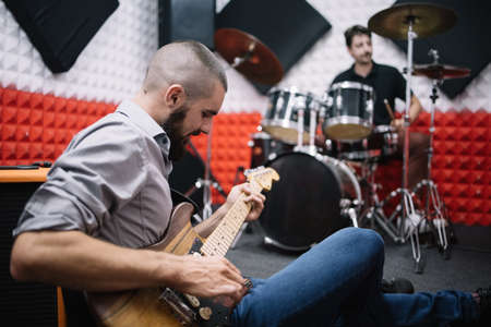 Guitarist playing guitar while sitting on floor in music studio. Electric guitar player sitting on ground next to drummer behind the drum set.の写真素材
