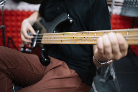 Close-up view of male hands playing electric guitar. Cropped male guitarist playing bass guitar while sitting in soundproof room.の写真素材