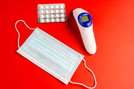 Pills, mask and digital thermometer on red surface. Virus accessories of mask, infrared thermometer...