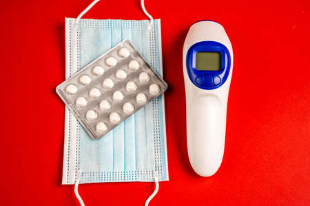 Top view of disposable mask and digital thermometer on red surface. Infrared thermometer, pills and face mask isolated on red backgroundの写真素材