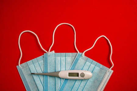 Top view of cropped surgical masks with digital thermometer. Digital thermometer over three disposable face masks isolated on red background.の写真素材