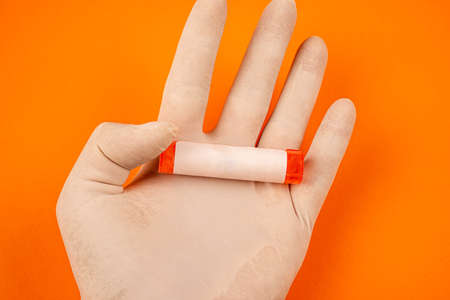 Top view of hand with white protective gloves holding vial with empty label. Cropped human hand with gloves holding small medical container over orange background.の写真素材